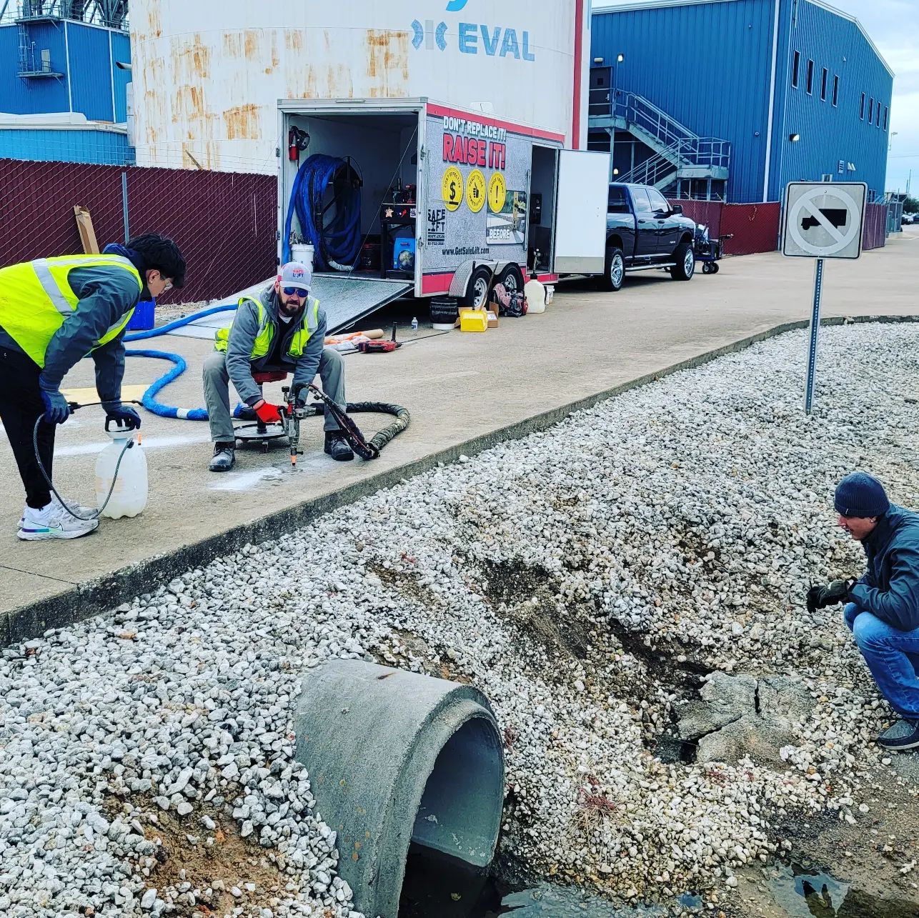 Three Men In Yellow Vests Are Repairing A Sewer Pipe, Focused On Their Work In An Urban Environment.
